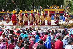 Audience and monks with ceremonial headdresses watching a spiritual dance.