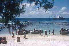 Carrying bags with supplies from the dinghy on the beach of Bakaka,  with the ship “Nei Momi” anchored offshore.
